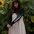 Woman in a garden with sunflowers wearing a dress and blacklong-sleeved t-shirt with floral print sleeves.