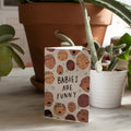 greeting card with text reading "babies are funny" encircled by cartoon babies with different skin tones and facial expressions and pale blue stars sitting infront of potted plants