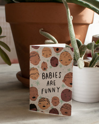 greeting card with text reading "babies are funny" encircled by cartoon babies with different skin tones and facial expressions and pale blue stars sitting infront of potted plants